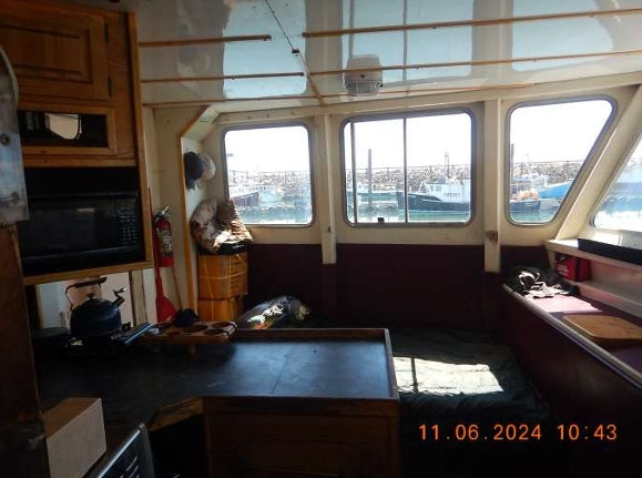 The interior of a boat cabin with wooden cabinets, a microwave, a table in the foreground, and large windows looking out onto a harbor. A timestamp is visible in the bottom right corner.