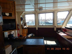 The interior of a boat cabin with wooden cabinets, a microwave, a table in the foreground, and large windows looking out onto a harbor. A timestamp is visible in the bottom right corner.