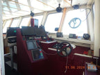 The helm station of a boat with deep red upholstered consoles, multiple electronic navigation screens, a black steering wheel, and large windows showing bright daylight.