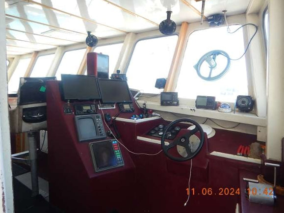 The helm station of a boat with deep red upholstered consoles, multiple electronic navigation screens, a black steering wheel, and large windows showing bright daylight.