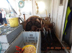 A large industrial mechanical unit with a wheel and hoses sits inside a cluttered workshop or storage area. In the foreground, a white crate holds a yellow bucket filled with small beige items, with stacked metal cases to the left.