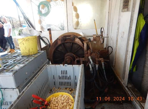 A large industrial mechanical unit with a wheel and hoses sits inside a cluttered workshop or storage area. In the foreground, a white crate holds a yellow bucket filled with small beige items, with stacked metal cases to the left.