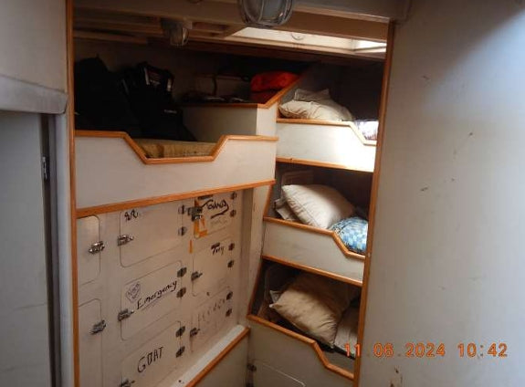 A narrow interior space with bunk-style beds and storage compartments, photographed inside a boat or cabin. The room has cream-colored walls and wood trim, with pillows and bags visible in the upper bunk area.