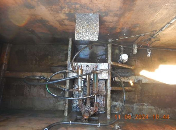A heavy-duty industrial hydraulic or mechanical control unit mounted on a rusty metal structure inside a confined industrial space. The unit features multiple black hydraulic hoses, metal cylinders, a diamond plate section, and wiring.