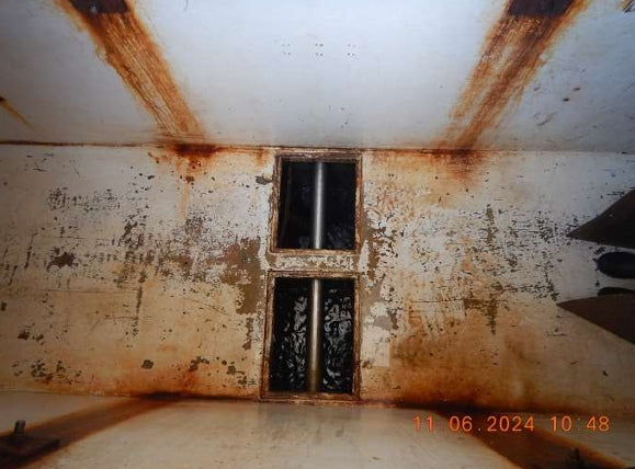 A dilapidated interior room with peeling off-white walls and ceiling, heavy rust stains, a barred window, and an orange timestamp in the lower right corner.