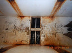 A dilapidated interior room with peeling off-white walls and ceiling, heavy rust stains, a barred window, and an orange timestamp in the lower right corner.