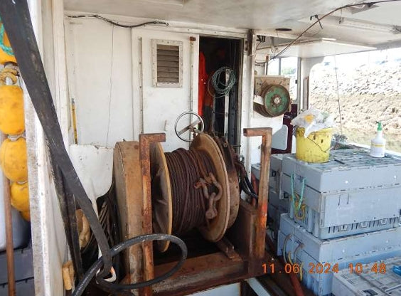A large wooden winch spool with coiled brown rope or cable, situated inside a boat cabin. To the left are yellow buoys, and to the right are stacked gray cooling crates.