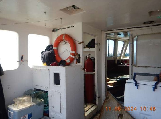 The interior of a boat cabin with white walls and windows, featuring an orange life preserver, a red fire extinguisher, a white cooler, and an open doorway looking into a darker navigation bridge. An orange timestamp is visible in the bottom right corner.