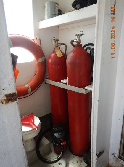Two red cylindrical gas cylinders mounted on a white metal rack inside a narrow storage compartment, with a life preserver ring on the left and a toilet visible below.
