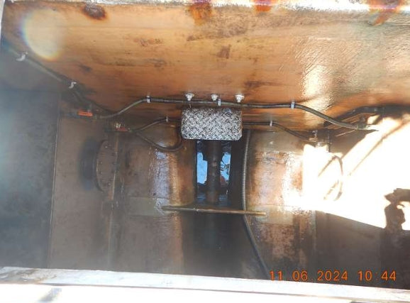 Close-up view of the underside of a rusty metal structure or machinery component, showing pipes, bolts, and a textured rectangular plate. A timestamp in orange appears in the bottom right corner.