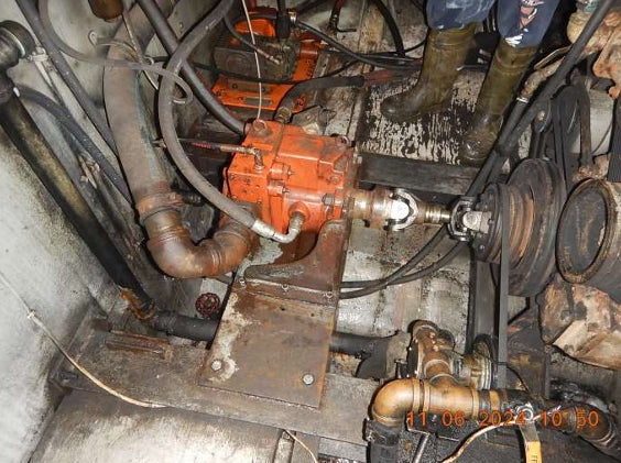 A used industrial mechanical transmission and pump assembly, featuring an orange painted metal housing, rusty metal pipes, black hoses, and a black pulley system, photographed in a workshop setting.