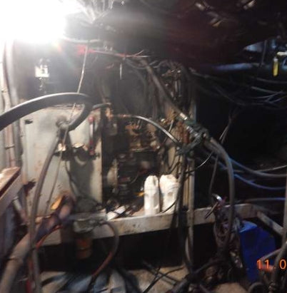 A close-up, cluttered view of an industrial machine or electrical compartment filled with cables, hoses, wires, and metal components, photographed in a dark, workshop-like environment with a bright glare at the top left.