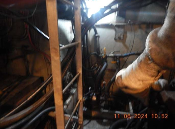A close-up, low-angle view of a mechanical system or furnace component in a dark, industrial setting, featuring a large insulated pipe, metal pipes, and a wooden ladder or support frame. A digital timestamp '11.