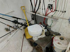 Small toilet mounted on a wall with fishing rods and various pipes and wires in a confined space.