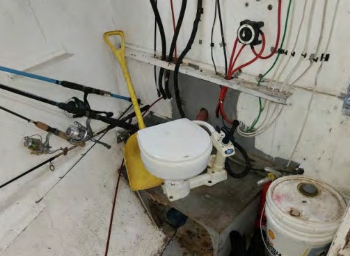 Small toilet mounted on a wall with fishing rods and various pipes and wires in a confined space.