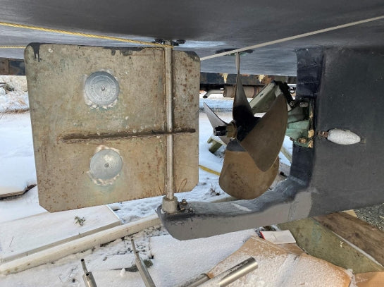 Close-up of a boat's stern mechanical area showing a large metal rudder, a bronze propeller, and part of the dark hull in a snowy outdoor setting.