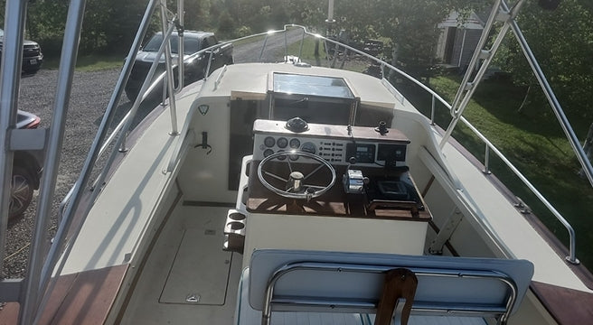 A rear-view perspective of a white motorboat with a wooden console, steering wheel, and navigation screens, docked outdoors on a gravel surface with green trees in the background.