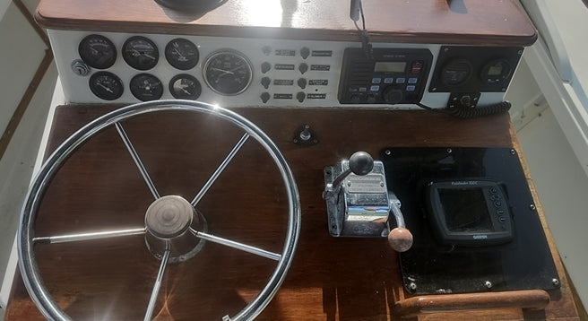 A close-up view of a boat's navigation console featuring a large chrome steering wheel, wooden dashboard, instrument gauges, a throttle lever, and a GPS screen.