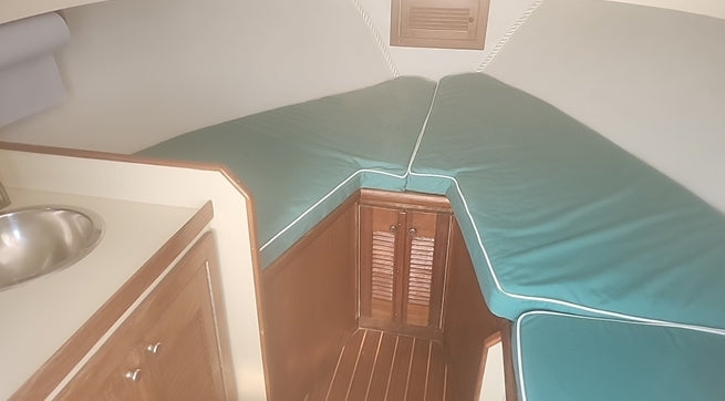 A close-up view of a boat cabin interior showing a teal green padded berth cushion with white piping, wooden cabinetry, a stainless steel sink, and a wooden slatted door.