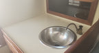 A stainless steel sink basin with a curved metal faucet installed in a cream-colored countertop, with a wooden-framed mirror above.