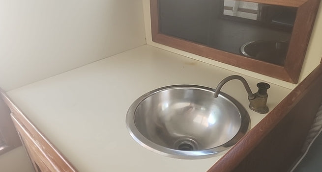 A stainless steel sink basin with a curved metal faucet installed in a cream-colored countertop, with a wooden-framed mirror above.