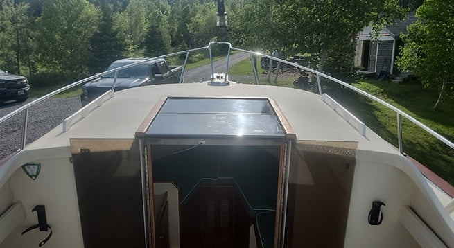 A top-down view from the deck of a boat, showing a glass windshield, bimbin, and the surrounding waterway with trees and a house in the background.