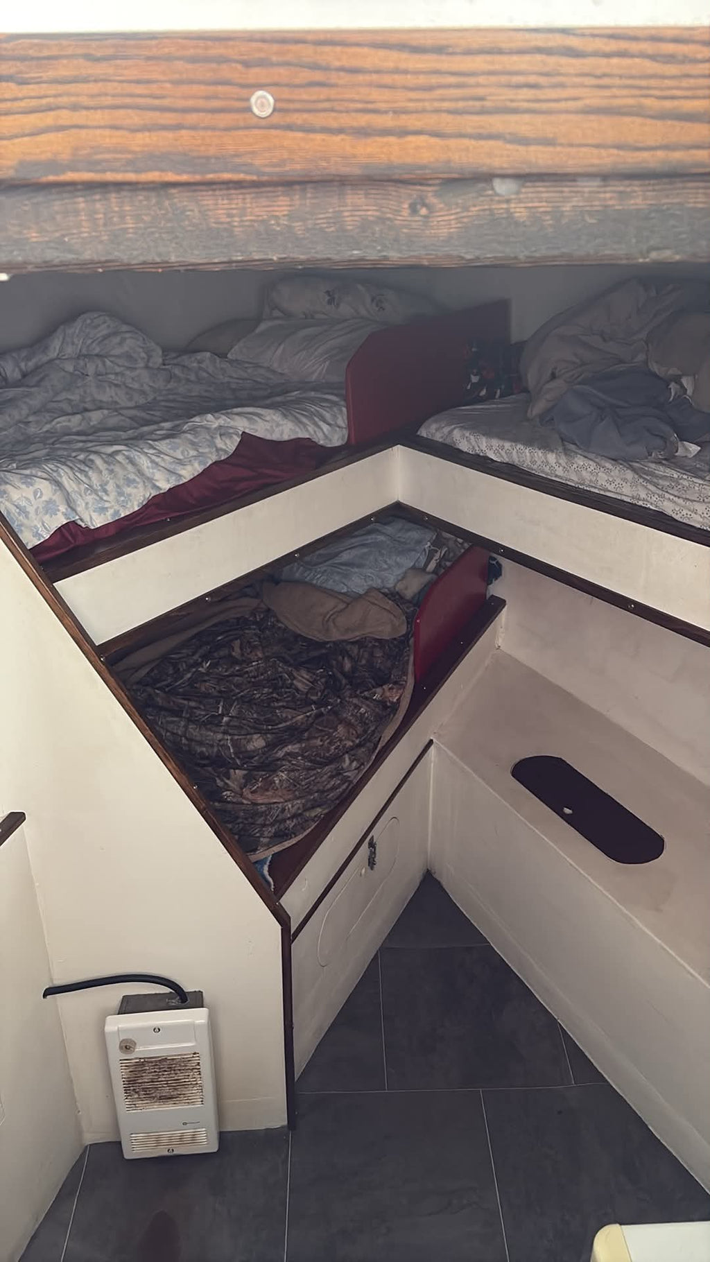 A compact L-shaped bunk bed setup inside a boat cabin, featuring two upper bunks with rumpled gray and white bedding, a lower bunk with dark patterned bedding, and a built-in storage bench on the right side.