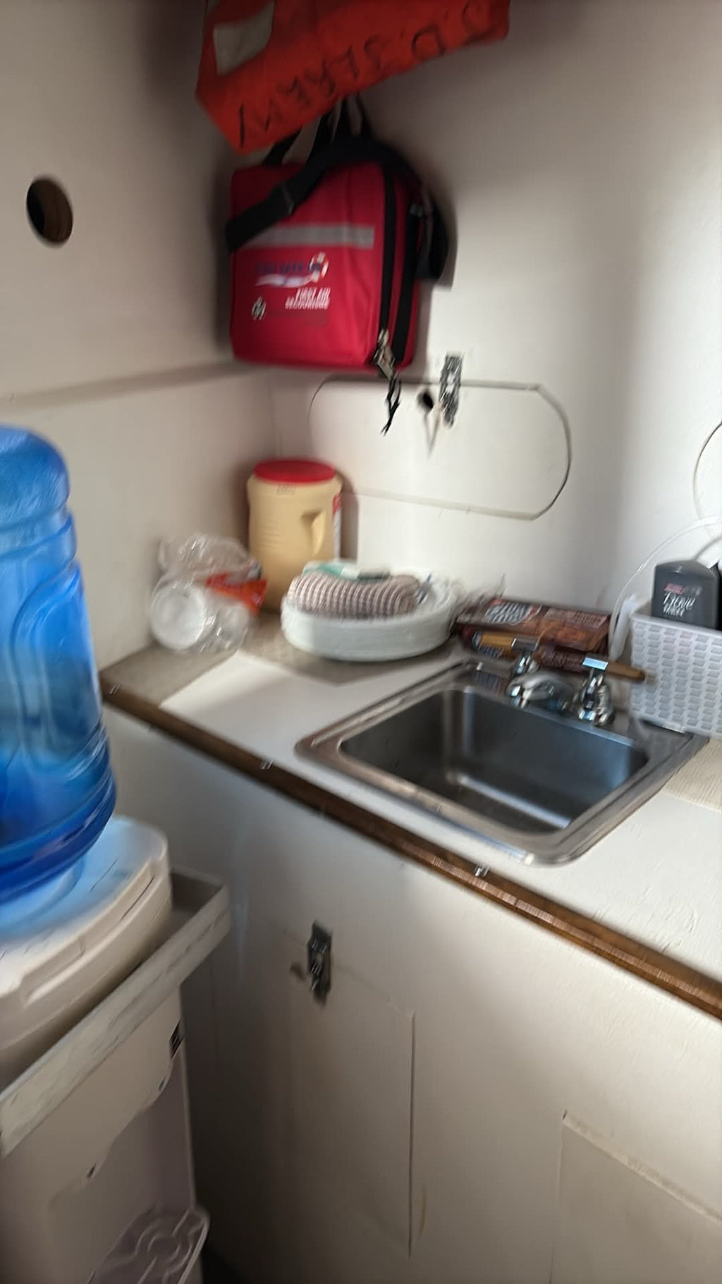 A compact kitchenette corner with a stainless steel sink, cream-colored cabinets, a countertop with dishes, and a red hanging bag on the wall.