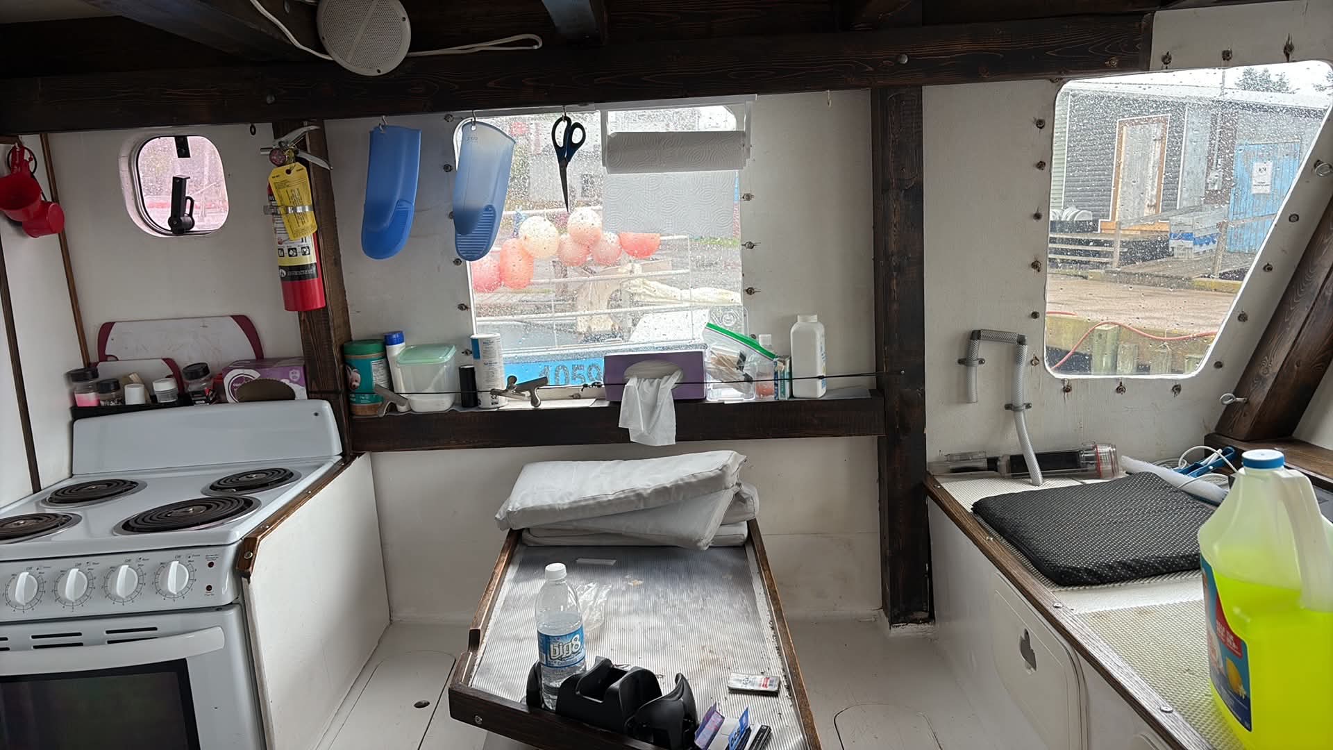 The interior of a boat cabin with a stove, galley sink, workbench, and windows looking out onto water and construction equipment.