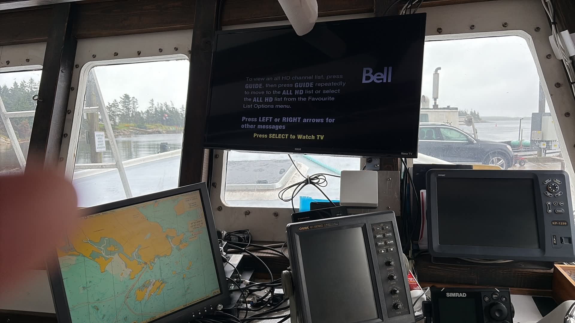 A cluttered boat bridge interior with multiple electronic screens, including a Bell-branded display and navigation devices, with water and trees visible through the windows.