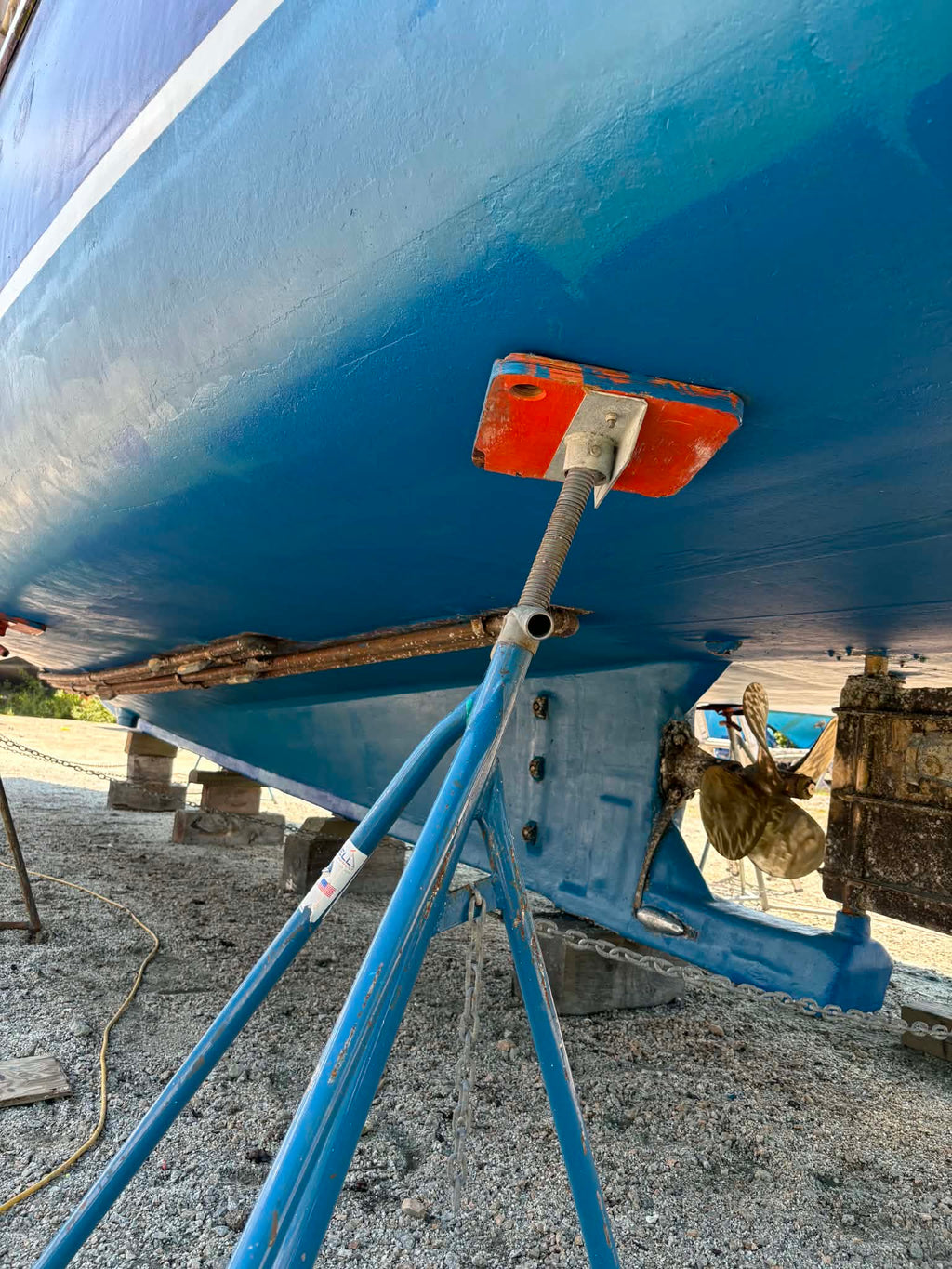 Close-up of the underside of a blue boat supported by metal stands on a gravel surface outdoors.