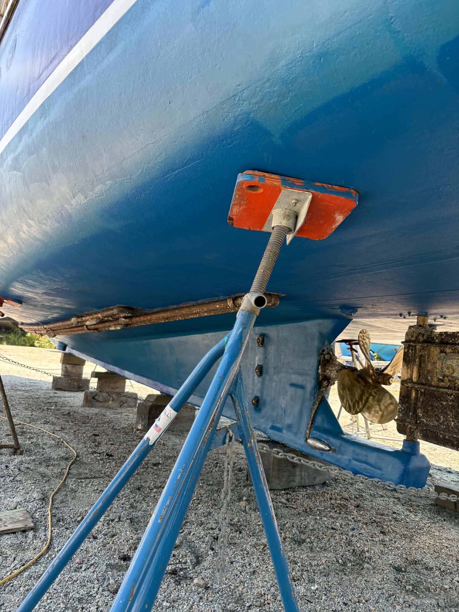 Close-up of the underside of a blue boat supported by metal stands on a gravel surface outdoors.