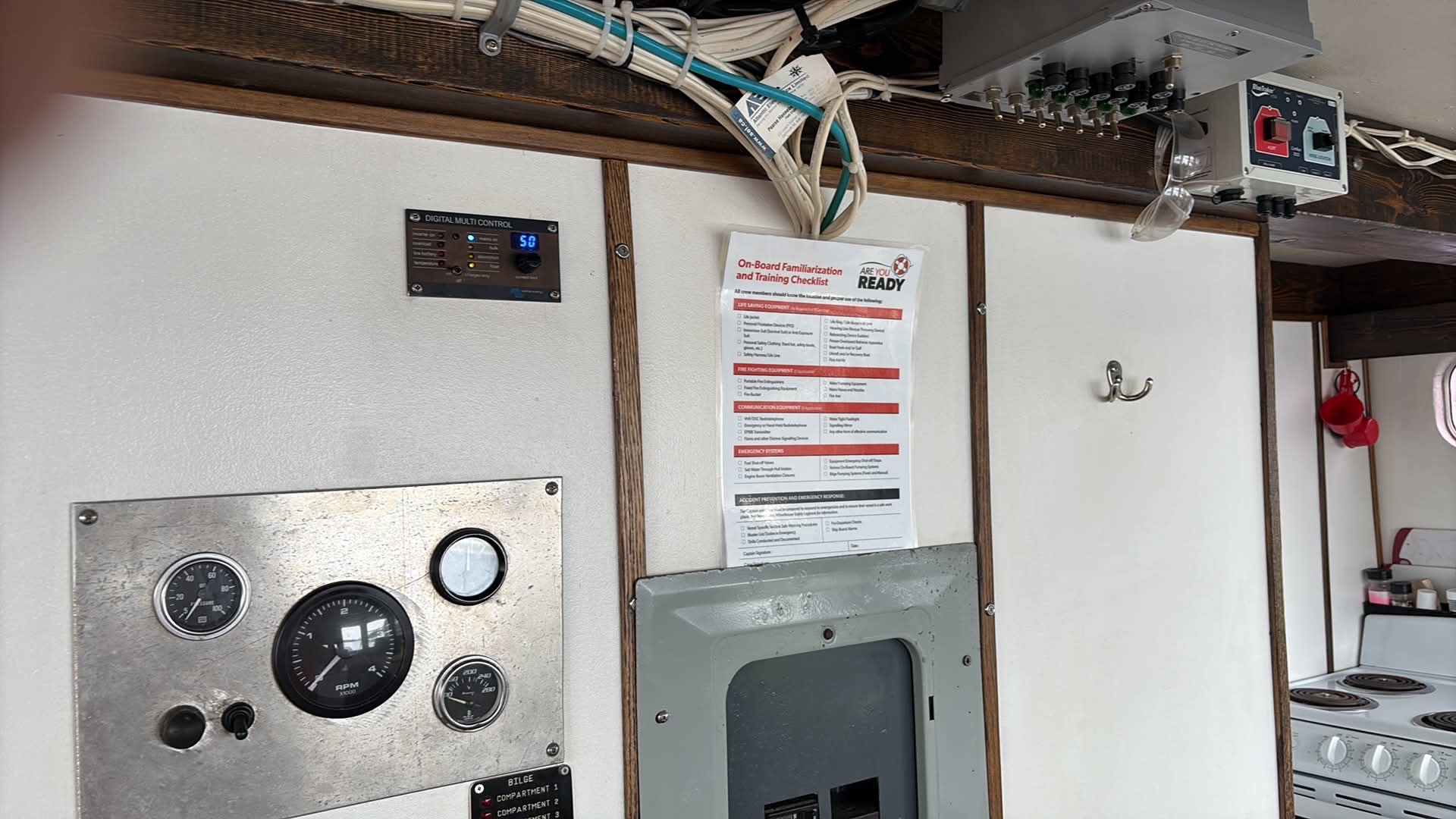An interior cabin wall with a metallic control panel, gauges, a hanging instruction sheet, electrical box, and wiring overhead on a boat.