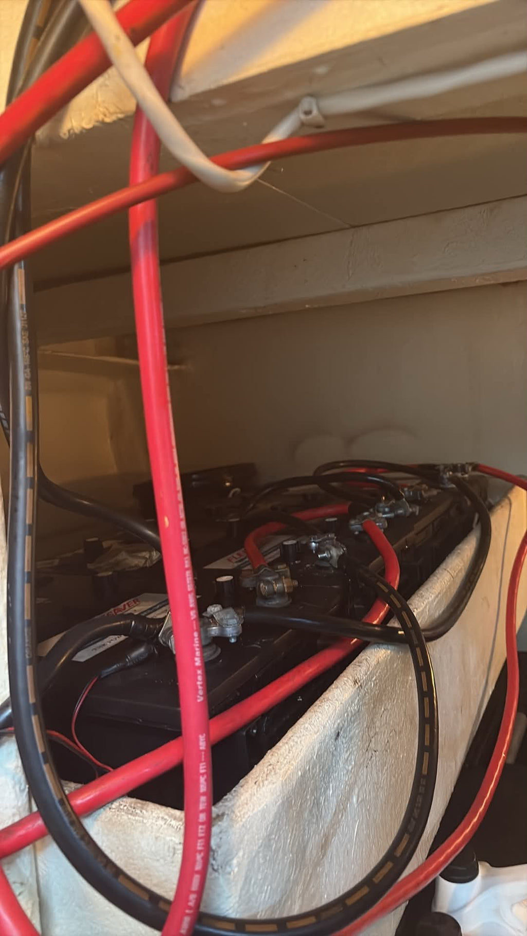A close-up view of a boat battery with thick red and black cables connected to its terminals, situated inside a boat cabin under a beige fiberglass console.