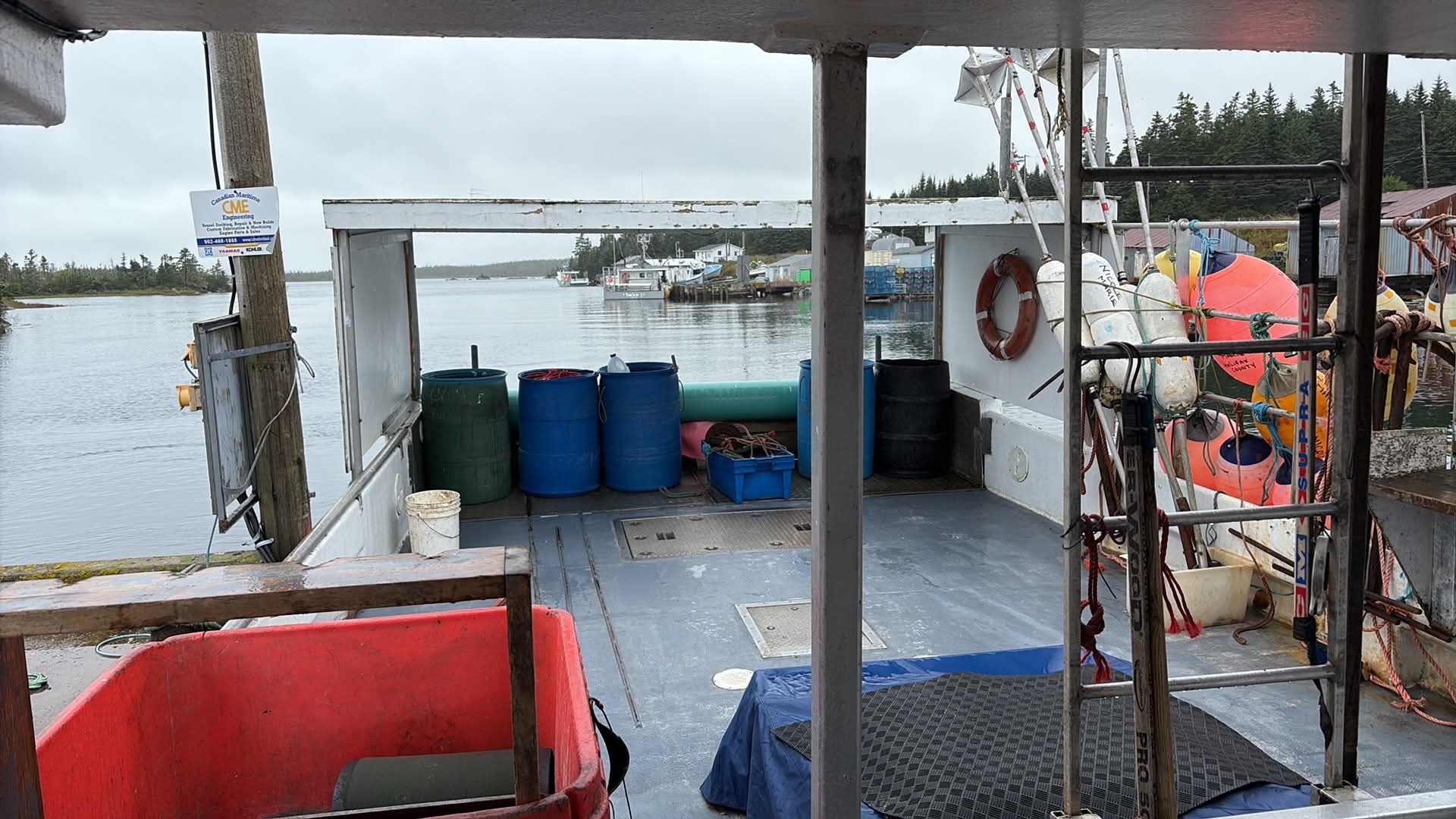 A view from the deck of a fishing boat in a harbor, with water, shoreline trees, blue barrels, life preserver rings, and fishing gear visible under an overcast sky.