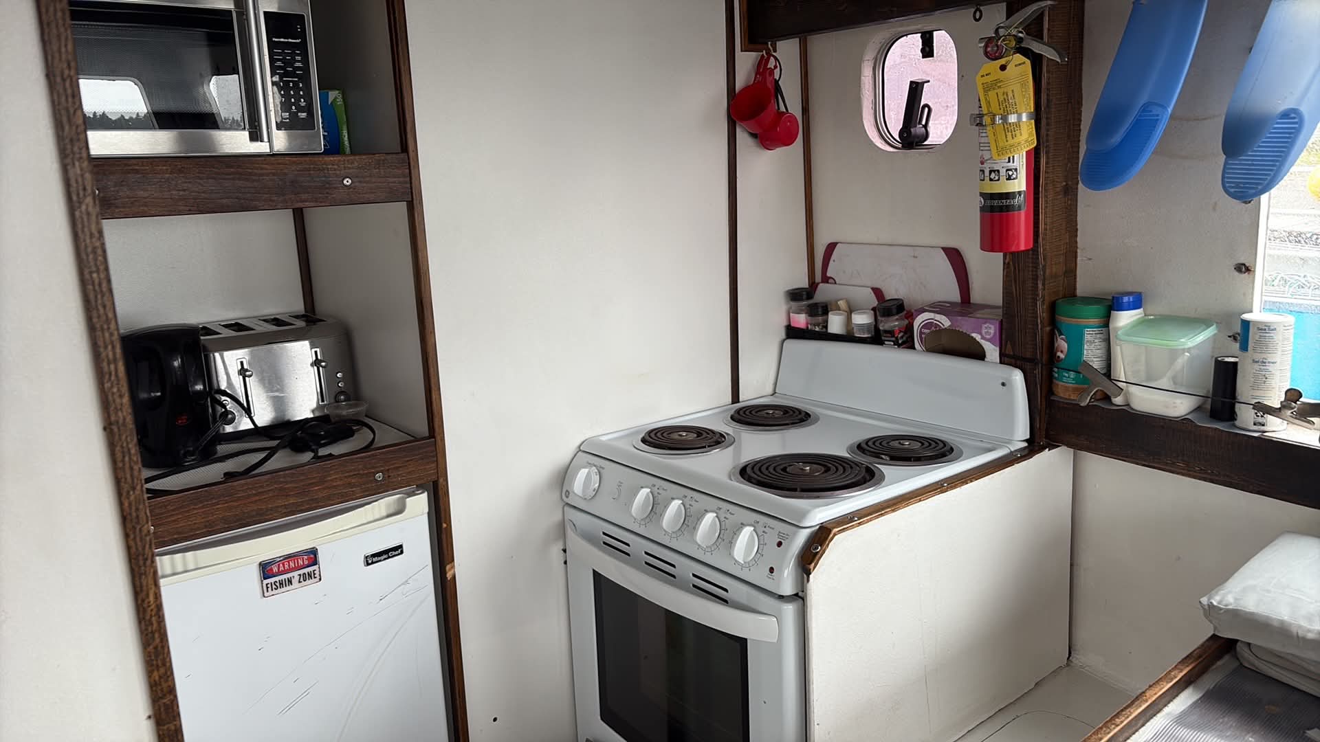 A compact white electric stove with coil burners in a small kitchenette setup, alongside a mini refrigerator, toaster, microwave, and hanging utensils.