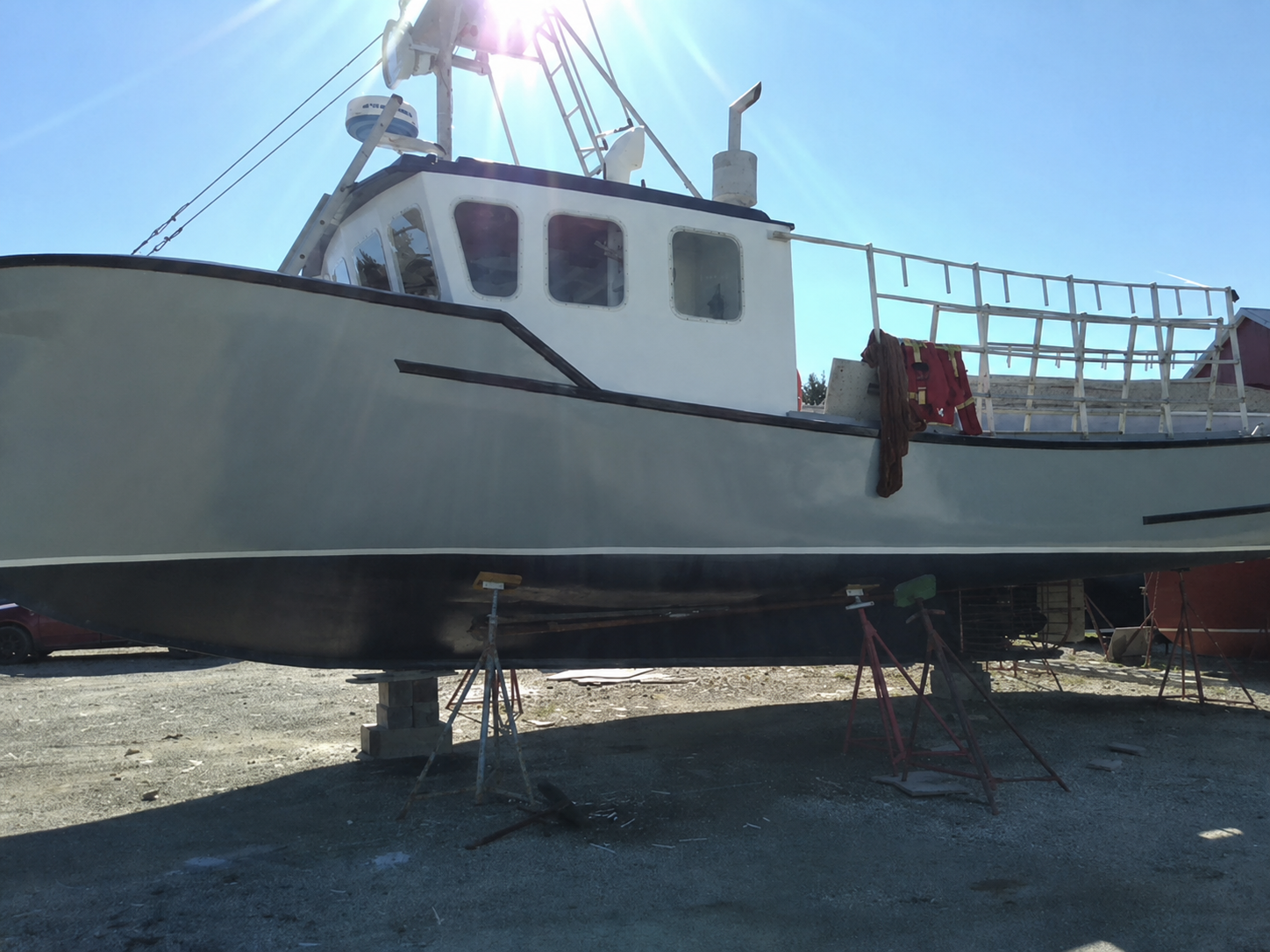 A large white and gray boat resting on metal support stands in an outdoor yard under a bright blue sky with sun glare.