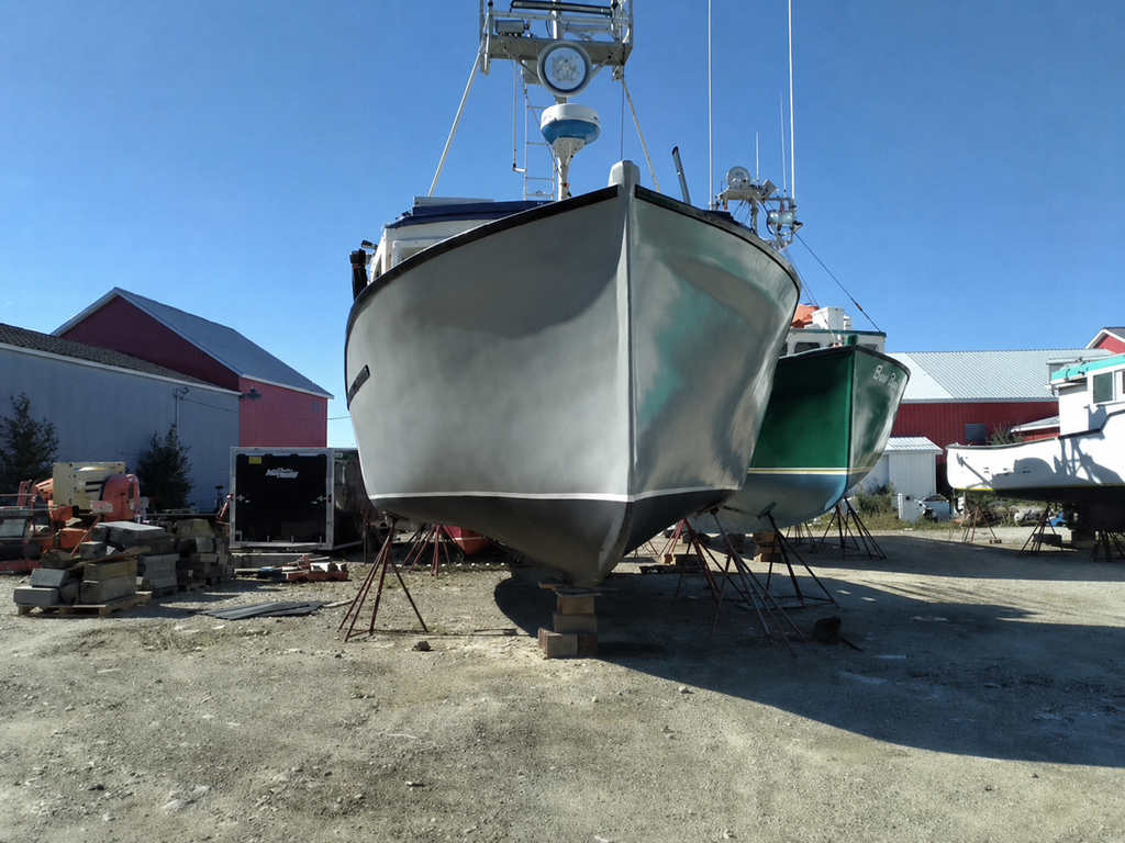 A gray motorboat on a lift in an outdoor boatyard, with other boats and red-roofed buildings visible in the background under a clear blue sky.