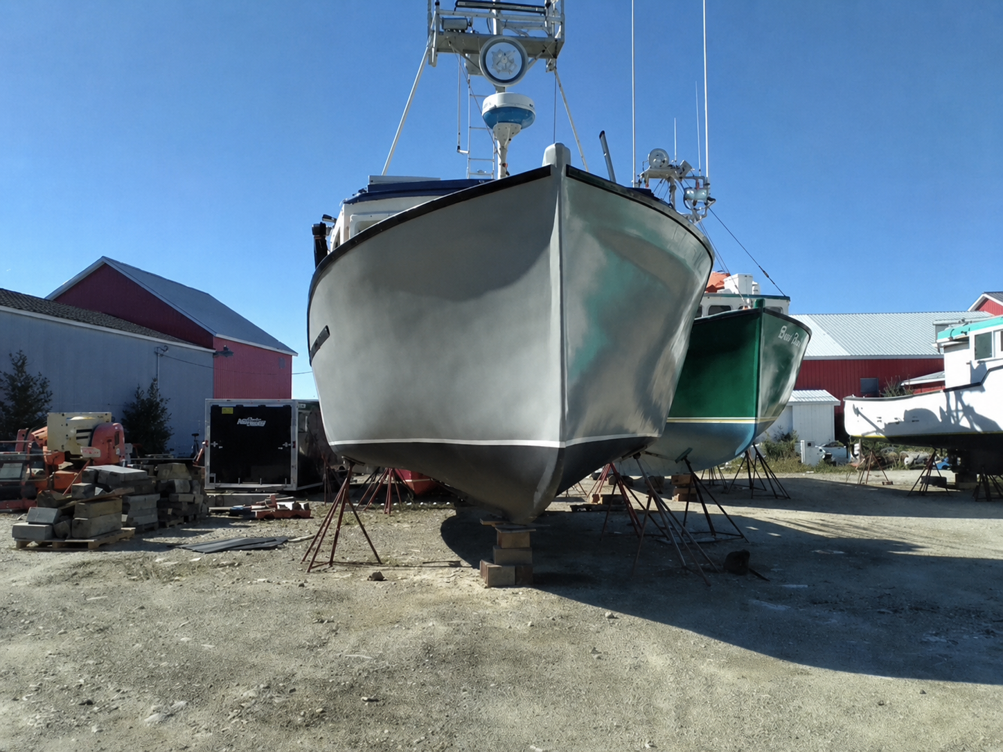 A gray motorboat on a lift in an outdoor boatyard, with other boats and red-roofed buildings visible in the background under a clear blue sky.