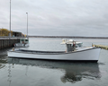 A white fishing boat with a dark hull is docked at a harbor under an overcast sky, with water, other boats, and a pier visible in the background.
