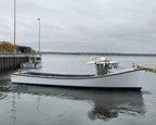 A white fishing boat with a dark hull is docked at a harbor under an overcast sky, with water, other boats, and a pier visible in the background.