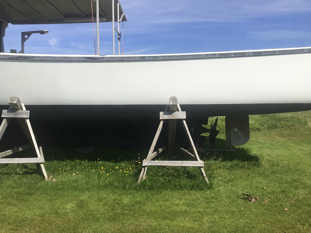 A white boat hull resting on wooden sawhorses outdoors on a grassy field under a blue sky.