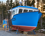 A blue and red boat on boat stands in an outdoor gravel lot surrounded by a forest at sunset.