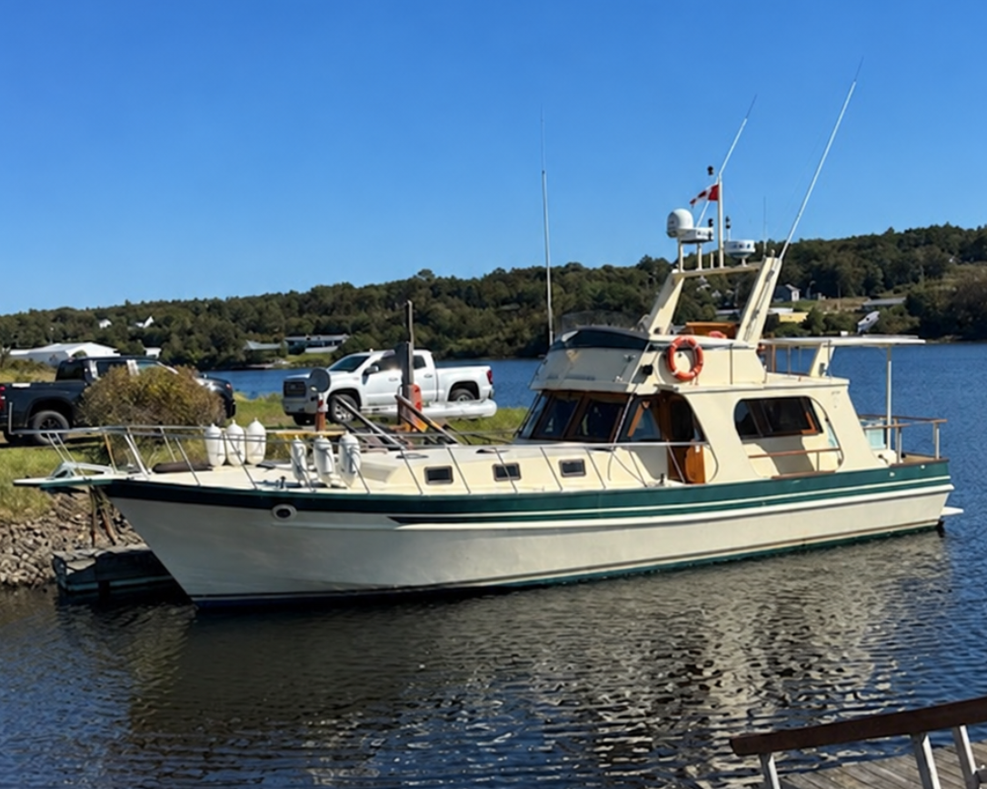 A cream-colored fishing boat with dark green stripes is docked on calm water under a clear blue sky, with a wooden dock in the foreground and trees in the background.