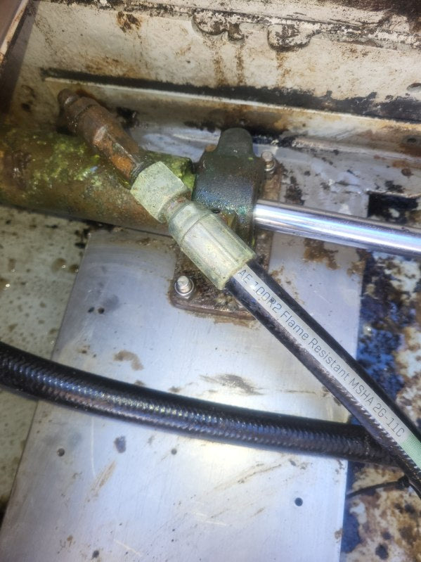 A close-up of a mechanical assembly showing a black flame retarder hose with printed text, a metal rod, and a brass fitting, set against a dirty, rusted metal background.