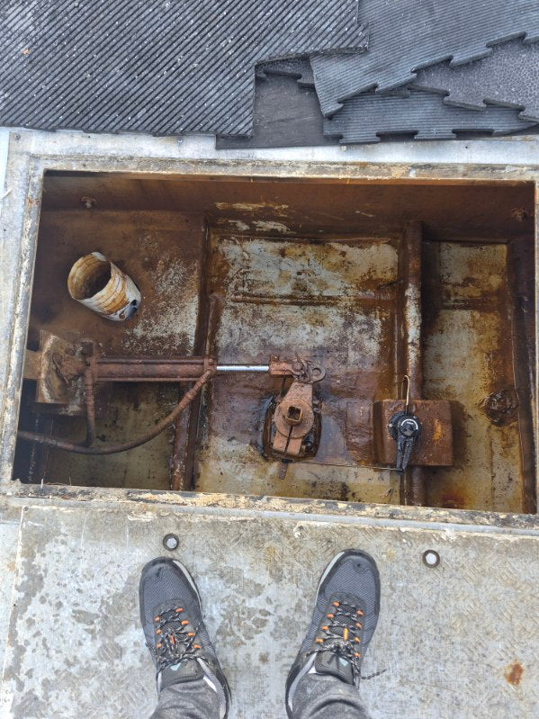 A top-down view of a rusty metal mechanical compartment or hatch opening, with a person's gray sneakers visible at the bottom edge on a concrete surface.