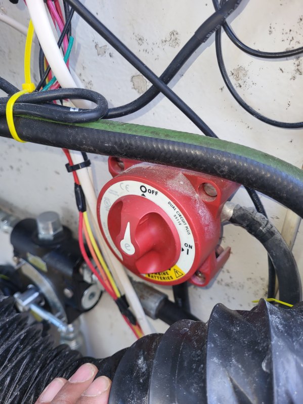 A close-up of a red electrical switch with a white dial and black cables connected to it, mounted against a light-colored wall with a hand visible at the bottom.
