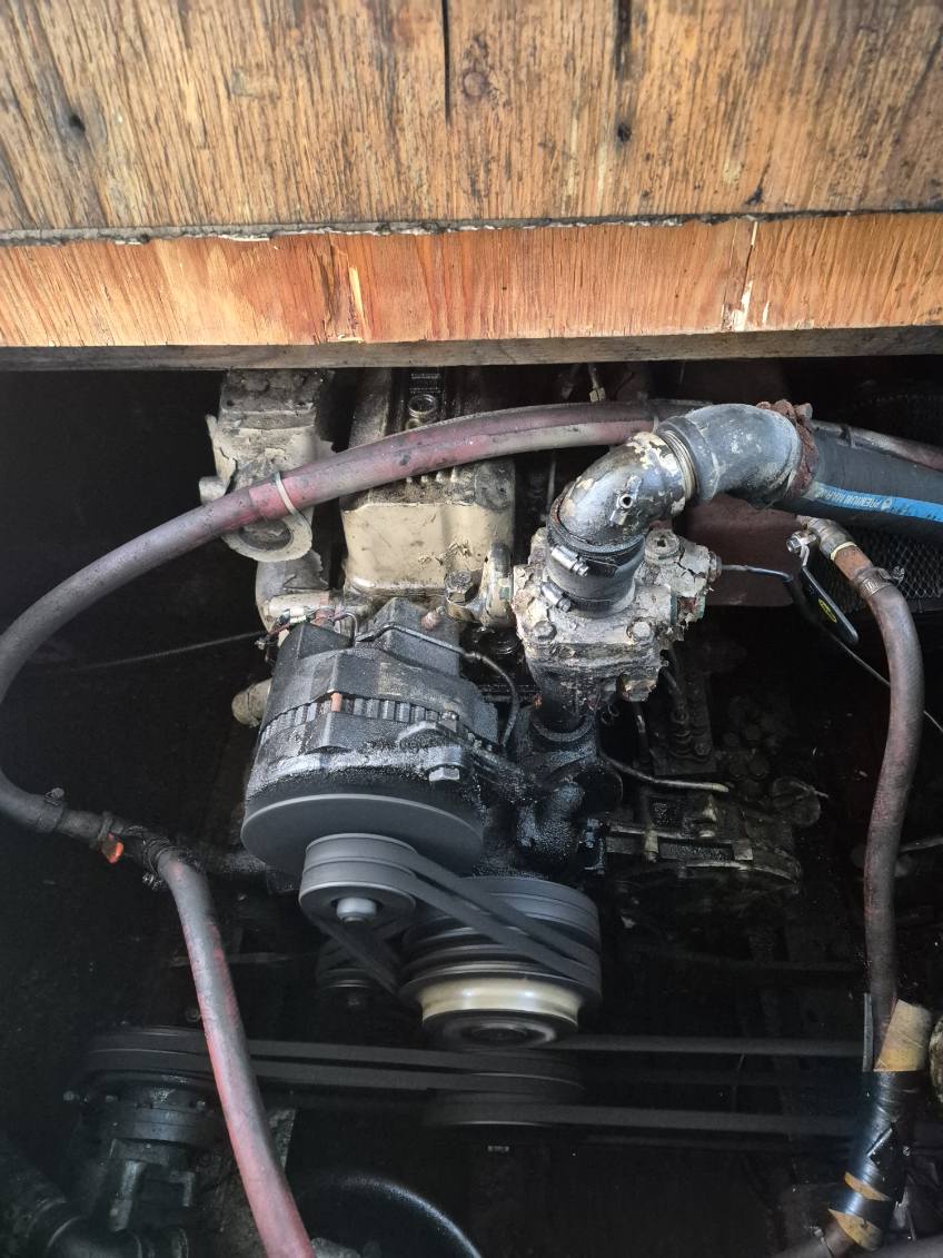 A close-up view of a used boat engine with a black alternator, pulley belt, and various hoses and metal components, situated under a wooden hull or deck structure.