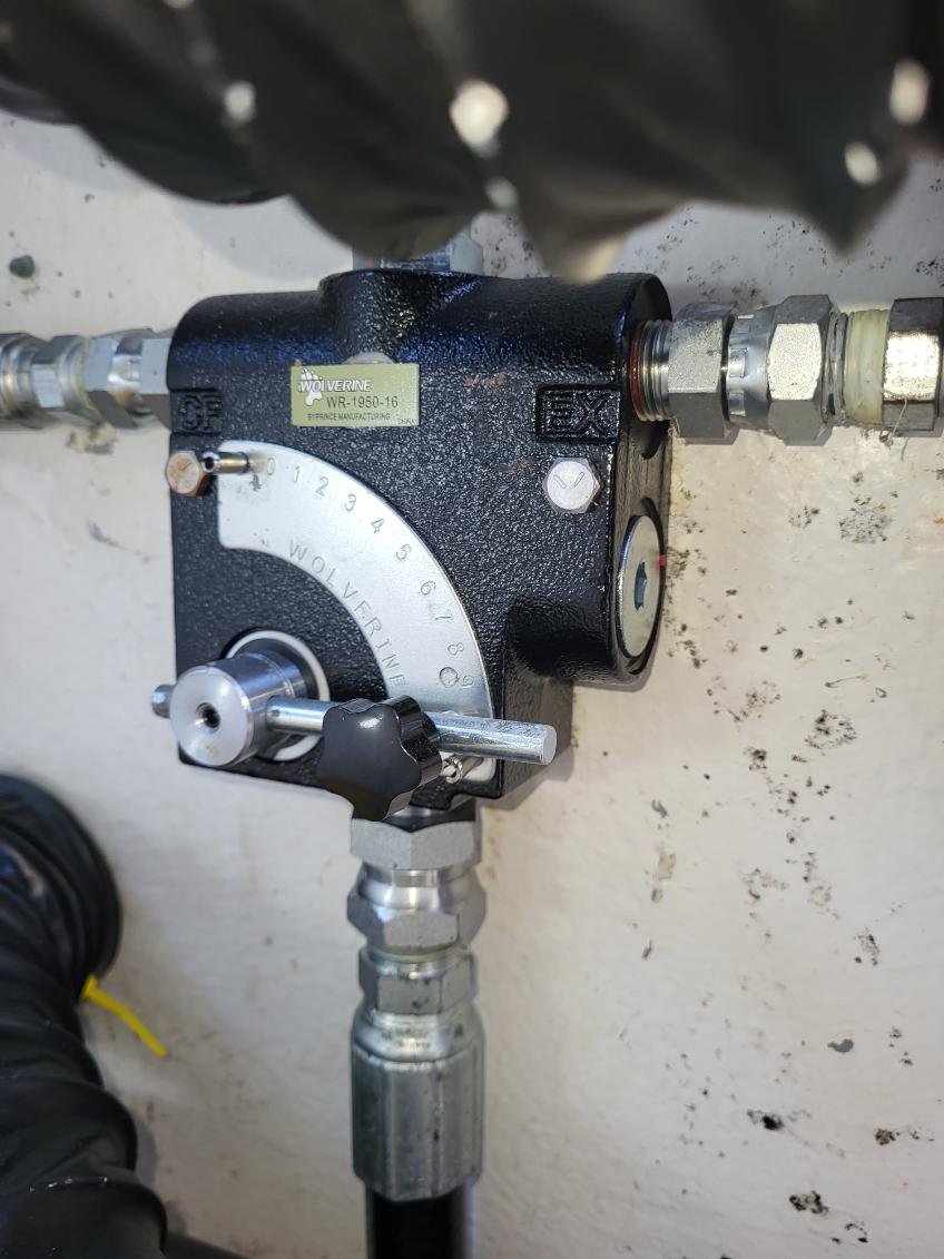 A close-up of a black industrial hydraulic or pneumatic control valve with metal fittings, mounted against a light concrete wall.