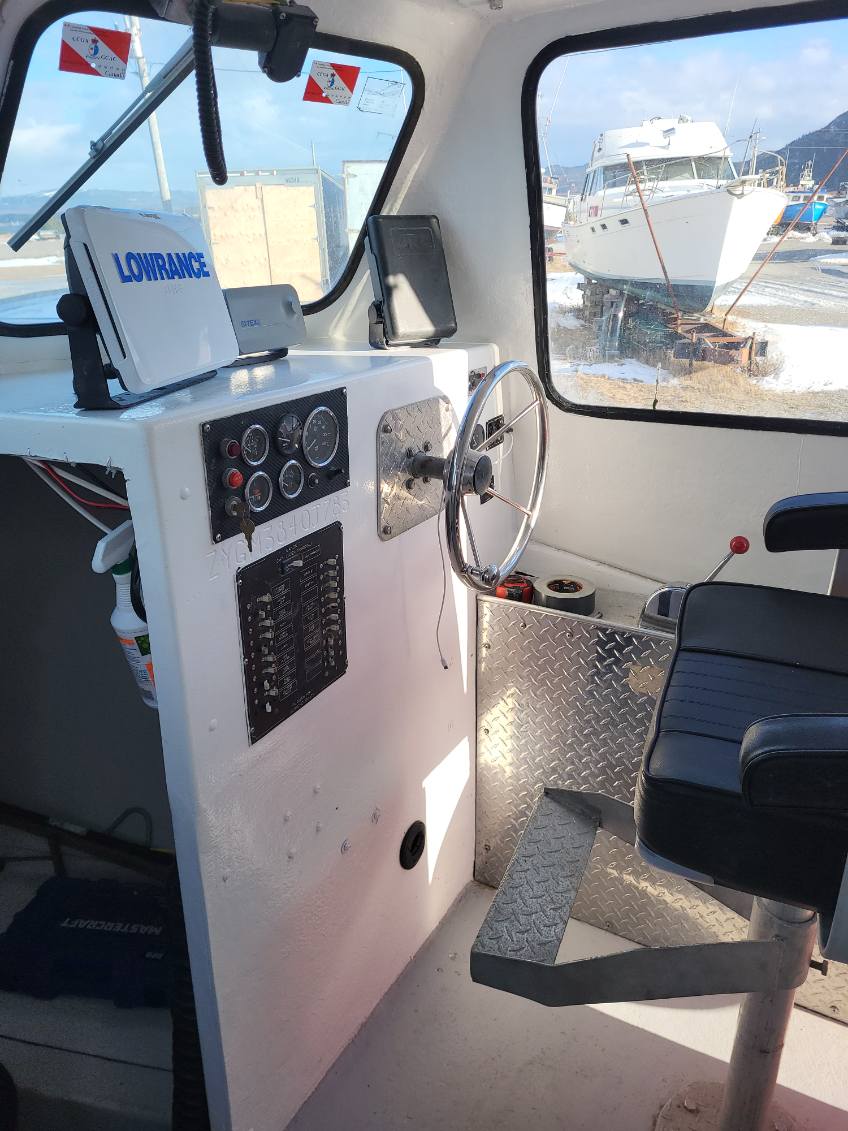 The helm station of a boat with a white console panel featuring a Lowrance GPS unit, steering wheel, and a black captain's seat. A boatyard scene is visible through the windows.
